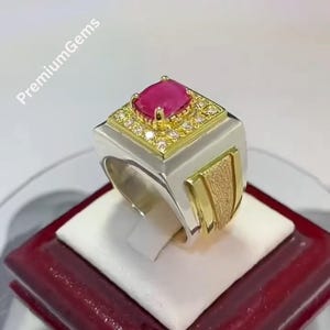 May include: A silver and gold ring featuring a large, square, pink gemstone. The gemstone is encircled by smaller, clear stones. The ring has a textured gold accent on the side. The text "Premium Gems" is visible.