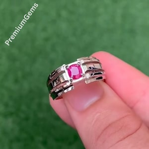 May include: A silver-toned metal ring featuring a central cushion-cut vibrant pink gemstone. The band has a multi-layered, stepped design on both sides of the stone. The text PremiumGems is visible in the upper left corner of the image.