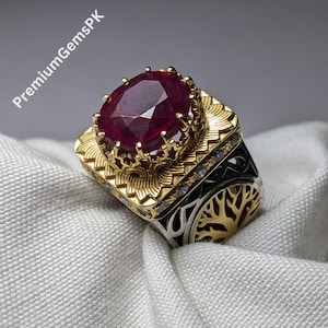 May include: A silver ring with a large red ruby gemstone set in a gold crown. The ring has a detailed design with a tree of life motif on the band.