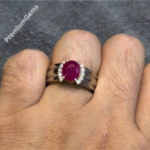 May include: A silver ring featuring a large, oval ruby gemstone, encircled by smaller, clear stones. The ring is displayed on a hand. The text "PremiumGems" is visible in the upper left corner.