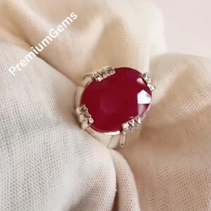 May include: A silver ring featuring a large, oval-cut, deep red gemstone. The ring has a detailed, claw-like setting around the stone. The text "PremiumGems" is visible in the upper left corner. The ring is displayed on a white fabric.