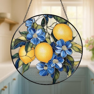 May include: Round stained glass window art featuring yellow lemons and blue flowers. The artwork is framed in black and suspended by a chain. The design includes green leaves and is set against a light background.