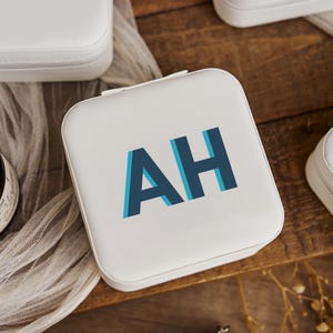 May include: White square travel case with the initials "AH" in blue and teal. The case is zippered and appears to be made of a smooth material. Other white cases are visible in the background, suggesting a set or collection.