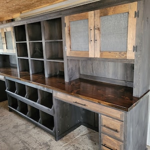 May include: A large, rustic wooden desk with a hutch. The desk has a dark wood finish and features a large, open shelf area, a set of drawers, and a hutch with two cabinets. The hutch has a wire mesh insert in the cabinet doors.