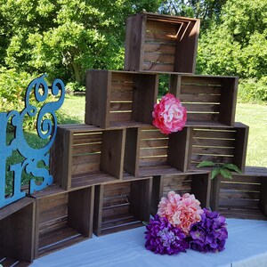 May include: A wedding display with wooden crates stacked to create a tiered structure, decorated with pink, purple, and green floral arrangements. A teal glitter sign reading "Mr & Mrs" is placed in front. The scene is set outdoors.