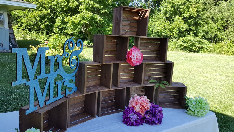 May include: A wedding display with wooden crates stacked to create a tiered structure, decorated with pink, purple, and green floral arrangements. A teal glitter sign reading "Mr & Mrs" is placed in front. The scene is set outdoors.