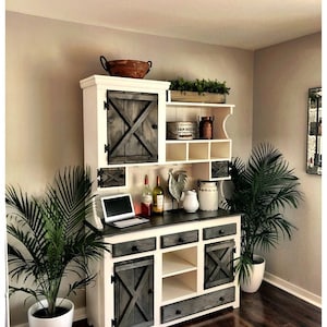 May include: A white and gray farmhouse style hutch with a distressed wood finish. The hutch has a top shelf with open shelves and a lower cabinet with drawers and doors. The doors have a cross-shaped design. The hutch is decorated with a rooster figurine, a pitcher, and a bowl.