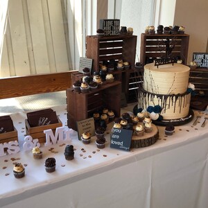 May include: A wedding dessert table featuring a two-tiered cake and cupcakes. The cake has light-coloured frosting with dark chocolate drips. Cupcakes are displayed on wooden crates and a wooden slice. Signs read "I love you more than cupcakes" and "you are loved."