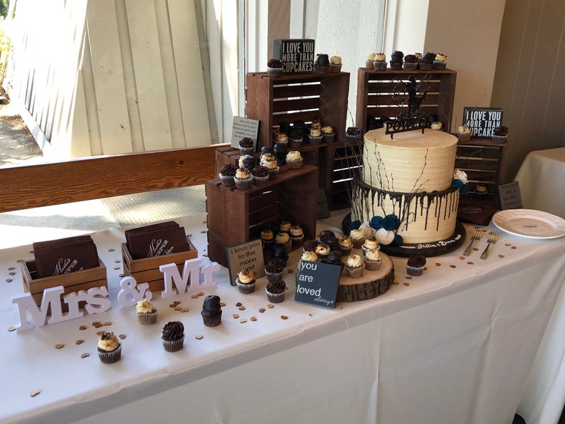 May include: A wedding dessert table featuring a two-tiered cake and cupcakes. The cake has light-coloured frosting with dark chocolate drips. Cupcakes are displayed on wooden crates and a wooden slice. Signs read "I love you more than cupcakes" and "you are loved."
