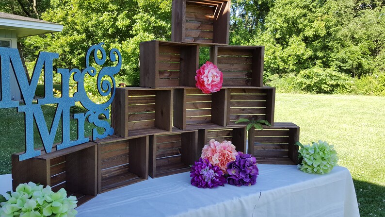 May include: A wooden crate display with a blue glitter "Mr & Mrs" sign. The display is decorated with colourful flowers, including pink, purple, and green, and is set on a white tablecloth.