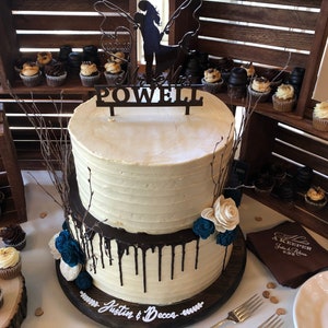 May include: A two-tiered wedding cake with white icing and chocolate ganache drips. The cake is decorated with blue and white flowers and a wooden topper that says "POWELL". Cupcakes and wooden crates are in the background.