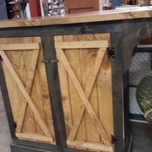 May include: A rustic wooden bar with a dark gray base and a light brown countertop. The bar features two wooden doors with a diagonal cross design. The doors are made of light-colored wood and are accented with black hinges. The bar has a classic, farmhouse style.