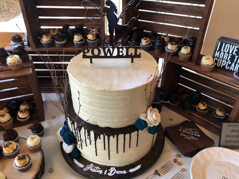 May include: A two-tiered wedding cake with white icing and chocolate ganache drips. The cake is decorated with blue and white flowers and a wooden topper that says "POWELL". Cupcakes and wooden crates are in the background.