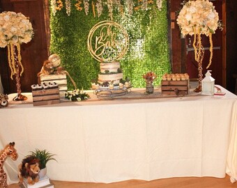 May include: A white table covered in a white tablecloth with a dessert table set up for a baby shower. The table is decorated with a wooden sign that says "Oh Baby" and a cake with a safari theme. There are also cupcakes, cookies, and other treats on the table.