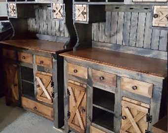 May include: Two rustic wooden hutch cabinets with a distressed grey finish and natural wood accents. Each cabinet features a top shelf with open cubbies, a closed cabinet door with a wood X design, and a lower cabinet with drawers and doors. The cabinets are displayed on a concrete floor.