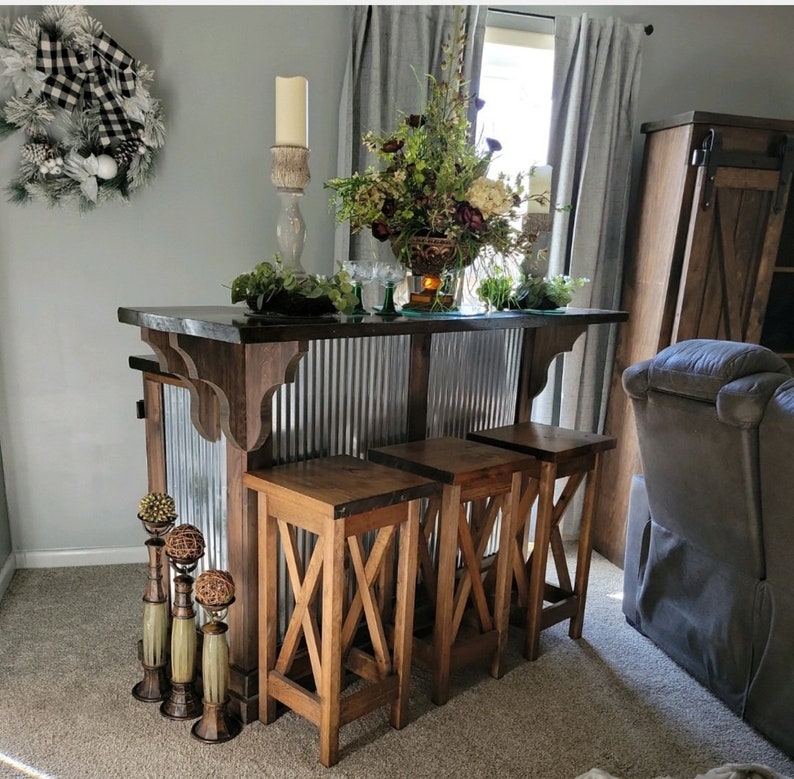May include: A rustic bar cart with a metal backsplash and three wooden stools. The bar cart has a dark wood finish and the stools have a natural wood finish. The bar cart is decorated with greenery and candles.