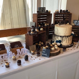 May include: A wedding dessert table featuring a two-tiered cake and cupcakes. The cake has light-coloured frosting with dark chocolate drips. Cupcakes are displayed on wooden crates and a wooden slice. Signs read "I love you more than cupcakes".
