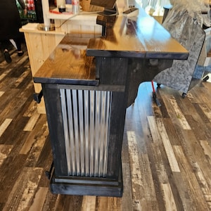May include: A black wooden bar counter with a dark wood top and a corrugated metal panel on the front. The counter has a shelf underneath and is sitting on a wood floor.