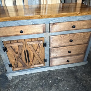 May include: A wooden cabinet with a stained top and a gray-painted frame. It features two drawers on top, three drawers on the right, and two barn-style doors. The cabinet has black hardware and a rustic design.