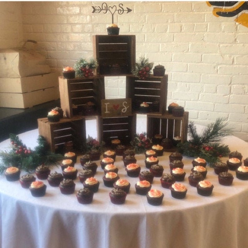 May include: A wedding cupcake display with a wooden crate tiered stand. The top crate has a sign that says "I & S" with an arrow pointing to the right. The cupcakes are frosted with red and orange frosting and are arranged on a white tablecloth.