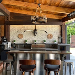 May include: Outdoor kitchen with a rustic wooden ceiling, a brick wall with a longhorn skull, a stainless steel grill, and a bar with metal siding and wooden stools.