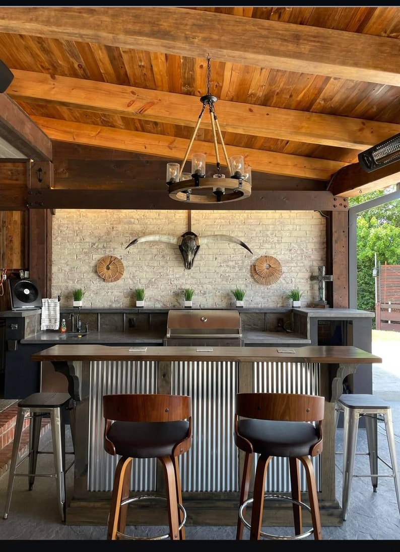 May include: Outdoor kitchen with a rustic wooden ceiling, a brick wall with a longhorn skull, a stainless steel grill, and a bar with metal siding and wooden stools.