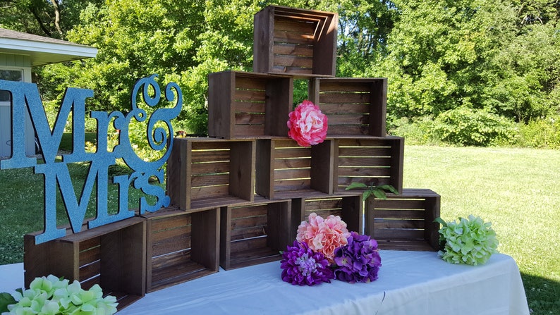 May include: A display of stacked wooden crates, forming a tiered structure. A large teal glitter sign reading "Mr & Mrs" is on the left. The display is decorated with colourful artificial flowers, including pink, purple, and green.