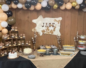 May include: A party table decorated with balloons, cupcakes, cakes, and food displays. A sign that says "JAGE" is displayed on the wall. The table is covered with a black and tan tablecloth.
