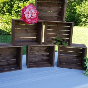 May include: Six dark brown wooden crates are stacked in a pyramid formation. A large pink and white flower sits atop the crates. The crates are on a white surface with a green backdrop of trees.