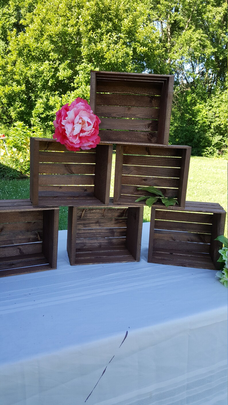 May include: Six dark brown wooden crates are stacked in a pyramid formation. A large pink and white flower sits atop the crates. The crates are on a white surface with a green backdrop of trees.