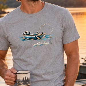 May include: A heather gray short-sleeve t-shirt featuring a graphic of a person wearing a hat and relaxing in a small boat while fishing with a rod. Below the boat, the text "au fil de l'eau" is printed in a cursive script. The shirt is made of cotton.