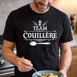Könnte beinhalten: Schwarzes T-Shirt mit dem weißen Text "TEAM COUILLÈRE" und einer Löffelgrafik. Das Design umfasst gekreuzte Schwerter und einen Helm über dem Text. Das T-Shirt ist kurzärmlig und aus dunklem Stoff.