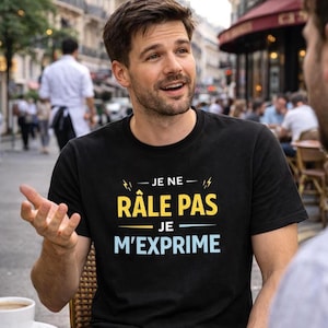Può includere: T-shirt nera girocollo con la scritta francese "JE NE RÂLE PAS JE M'EXPRIME" in giallo e azzurro. La t-shirt è indossata da una persona in un ambiente esterno.