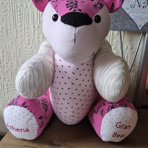 May include: A handmade pink and white teddy bear with floral and heart patterns. The bear has black button eyes and a black nose. The bear's paws are inscribed with the names "Athena" and "Gran Bear".