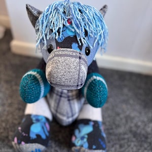 May include: A stuffed horse toy crafted from various fabric patterns, including a grey plaid muzzle, a black fabric with blue character prints, and a solid grey section. It features a bright blue yarn mane and two textured teal fabric hooves.