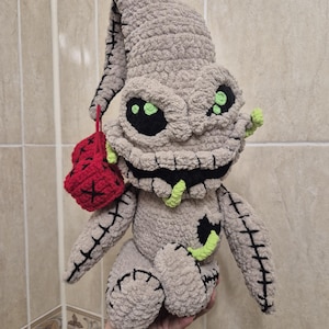 Op de afbeelding: Een gehaakt pluche speelgoed dat lijkt op een personage uit de film "The Nightmare Before Christmas". Het speelgoed is grijs met groene ogen en een gestikte mond. Het houdt een rode gehaakte tas vast.