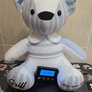 May include: A memory bear crafted from light blue and white fabric with striped and star patterns. The bear features black button eyes, a black nose, and a smocked chest panel. Text includes DUMBO, 2026, Griff, and 22/04/26 with 8.3lb. A digital scale shows 832.