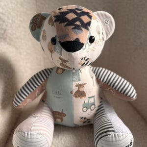 Puede incluir: Un oso de peluche hecho de varios parches de tela con estampados de granja, incluyendo tractores y vacas. El oso tiene ojos de botón negros, una nariz negra y extremidades a rayas. El pie izquierdo tiene bordado "Oscar" y el derecho "09/12/26".