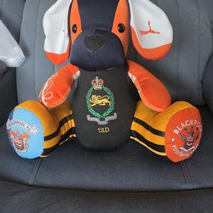 May include: A plush dog toy constructed from orange, black, and white fabric panels. The chest features a crest with a lion and the text 1.3.D. The feet display Blackpool F.C. logos. One ear has a white Puma logo, and the face has black eyes.