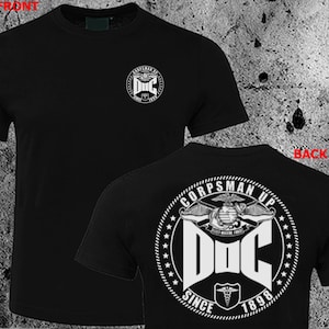 Puede incluir: Camiseta negra con gráficos blancos en la parte delantera y trasera. El gráfico frontal dice "CORPSMAN UP" y "DOC SINCE 1898". El gráfico trasero dice "CORPSMAN UP" y presenta un símbolo médico.