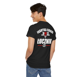 May include: Black t-shirt with white text reading "FABRYKA BRONI" above "ŁUCZNIK" and the number "11". The text includes the years "19 22" and the city "RADOM POLAND". The shirt is worn by a person.