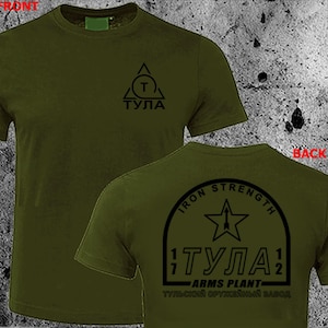 May include: Olive green t-shirt with black graphic designs. The front features a triangle with a 'T' inside and the word 'TULA'. The back displays a star within an arch, the words 'IRON STRENGTH', and the word 'TULA' again, along with 'ARMS PLANT'.