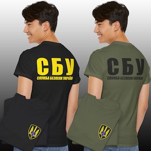 May include: Two short-sleeved t-shirts, one black and one olive green, each featuring the yellow or black Cyrillic text SBU and the Ukrainian Security Service emblem on the chest and back, displaying a shield with a sword and wings.