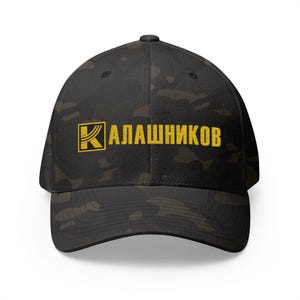 Op de afbeelding: Zwarte baseballcap met een camouflagepatroon. De cap heeft het woord "Калашников" in het geel, met een gele vierkant en een "K" in de linkerbovenhoek.