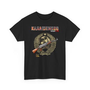 May include: Black t-shirt featuring a graphic of an AK-47 assault rifle, with the text "Kalashnikov" in red, white, and gold. The design includes a wheat wreath and a globe. The caliber is noted as 5.45.