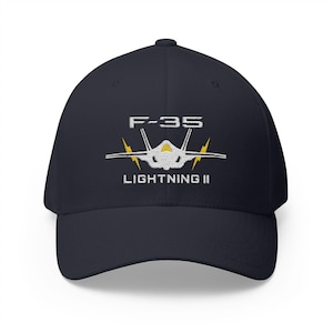 Peut inclure: Casquette de baseball bleu marine avec le texte "F-35 LIGHTNING II" brodé en blanc. Le design comprend un avion stylisé et des éclairs jaunes. Casquette classique et structurée.