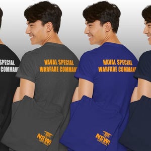 Op de afbeelding: Vier korte mouwen t-shirts in zwart, grijs, blauw en marineblauw. Elk shirt heeft de tekst "NAVAL SPECIAL WARFARE COMMAND" op de rug. De voorkant van elk shirt toont de tekst "NSWI" en een logo.