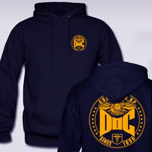 Puede incluir: Sudadera con capucha azul marino con bolsillo delantero y cordón. La parte delantera presenta un emblema dorado con el texto "CRAFTSMAN OF FINE ARTS SINCE 1898". La parte trasera muestra un emblema dorado más grande con el texto "GOT UP DOC SINCE 1898".