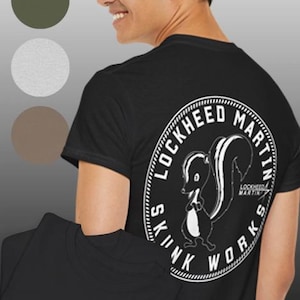 Könnte beinhalten: Schwarzes T-Shirt mit einem weißen Grafikdesign eines Stinktiers in einem Kreis mit dem Text "Lockheed Martin Skunk Works". Das Shirt hat auch eine kleinere Version der Grafik auf der Vorderseite.