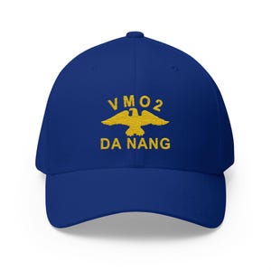 Op de afbeelding: Een koningsblauwe baseballcap met een gebogen klep. De cap heeft de tekst "VMO2 DA NANG" in gouden letters, met een gouden adembleem boven de stadsnaam. Een klassiek, casual accessoire.